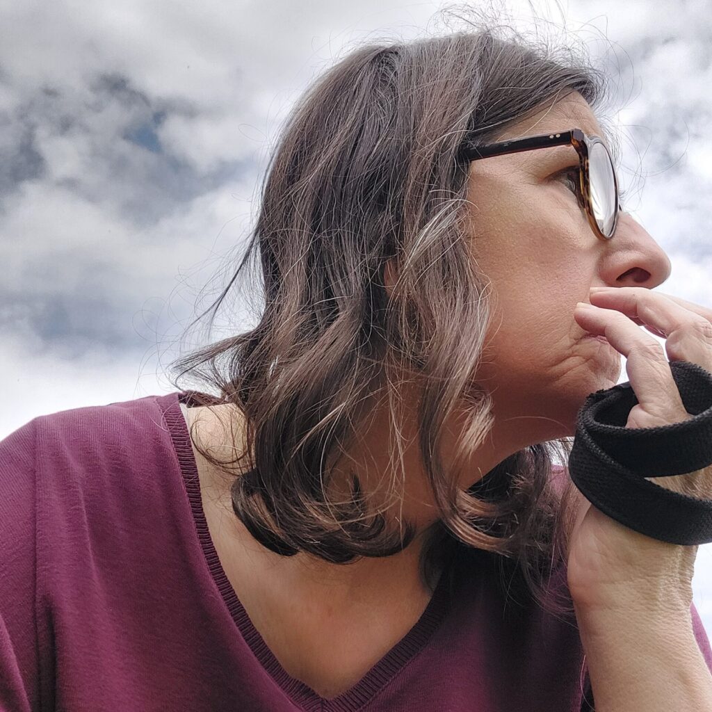 Nahaufnahme einer Frau mit Brille, die nachdenklich zur Seite blickt. Der Himmel im Hintergrund ist bedeckt. Ihr Gesichtsausdruck wirkt ruhig, aber ernst. Eine schwarze Hundeleine liegt locker um ihre Hand.