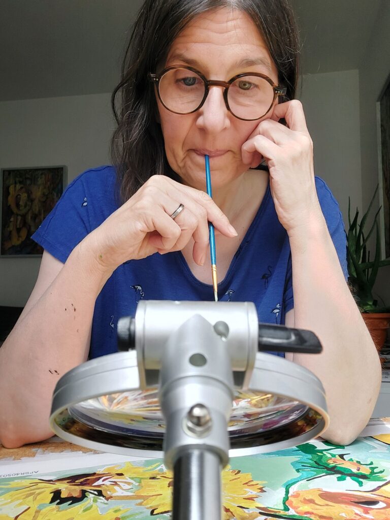 Frau mit Brille sitzt am Tisch und malt Van Goghs Sonnenblumen nach Zahlen. Nachdenklich hält sie den Pinsel im Mund, im Vordergrund eine Lupe.