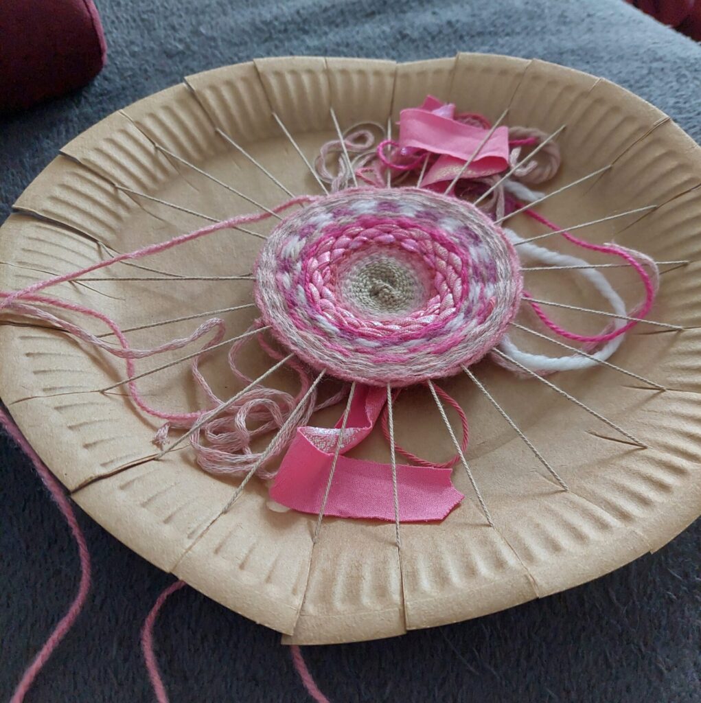 Einfacher Webrahmen aus einem eingeschnittenen Pappteller. Mit pinker und weißer Wolle entsteht ein rundes Webmuster, durchzogen von Bändern.
