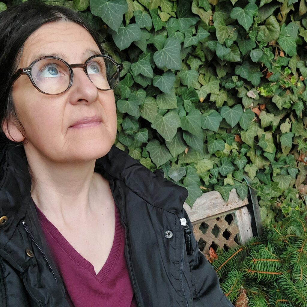 Frau mit Brille sitzt vor einer efeubewachsenen Wand und blickt nachdenklich nach oben. Herbstliche Stimmung, Laubarbeit liegt hinter ihr.