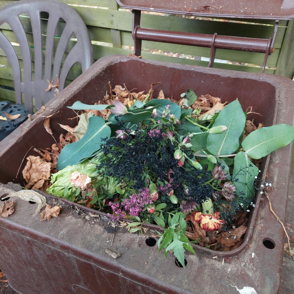 Verwelkte Blumen und Laub liegen in einer braunen Biotonne. Die Szene wirkt herbstlich und ruhig – Zeichen eines bewussten Loslassens.