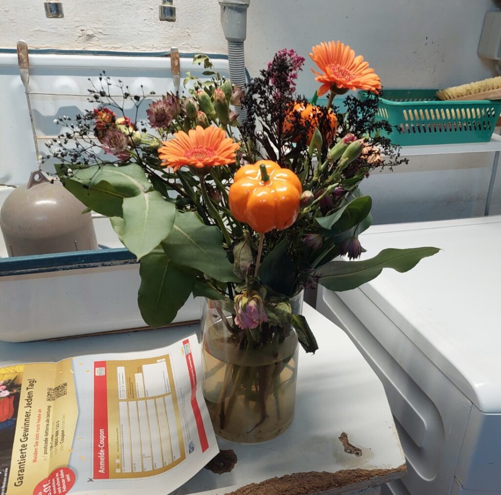 Ein verblühter Blumenstrauß mit orangefarbenen Gerbera und Dekokürbissen steht in einer Vase auf einer alten Arbeitsplatte. Einzelne Elemente werden gerettet.