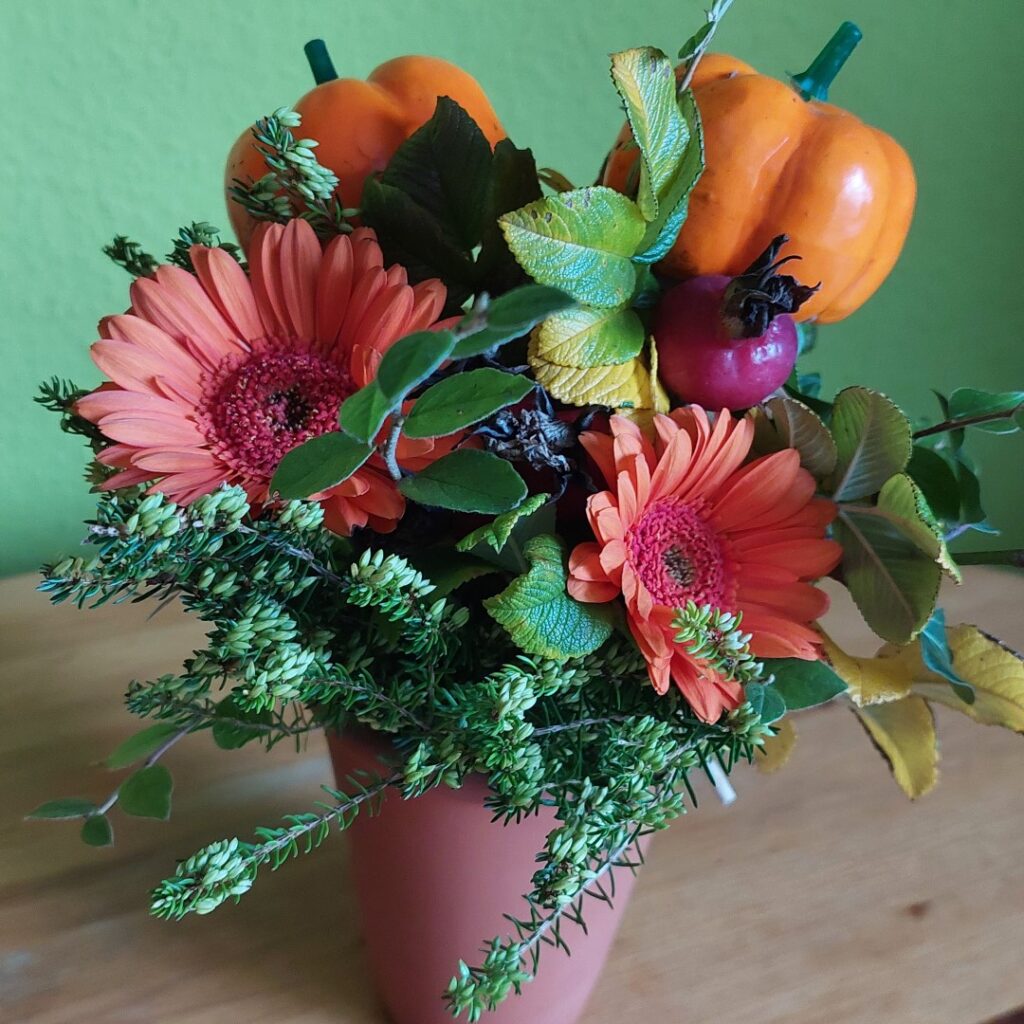 Ein frischer Strauß mit Gerbera, Zierkürbissen, Hagebutte und Gartenpflanzen steht in einer Vase vor grüner Wand. Eine kreative Neuschöpfung aus alten und neuen Teilen.