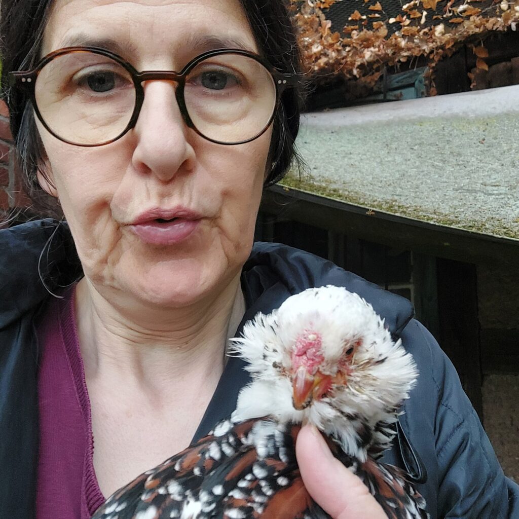 Frau mit Brille hält ein geschecktes Huhn auf dem Arm und schaut mit weichem Ausdruck. Das Huhn ist in der Mauser, neue Federn wachsen nach.