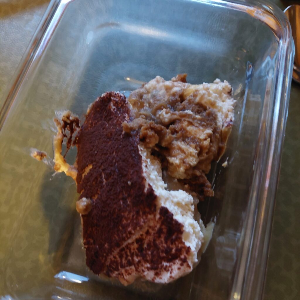 Glasbehälter mit einem Rest Kuchen oder Dessert. Zu sehen sind verschiedene Schichten, teilweise zerfallen, mit Creme und Teig.