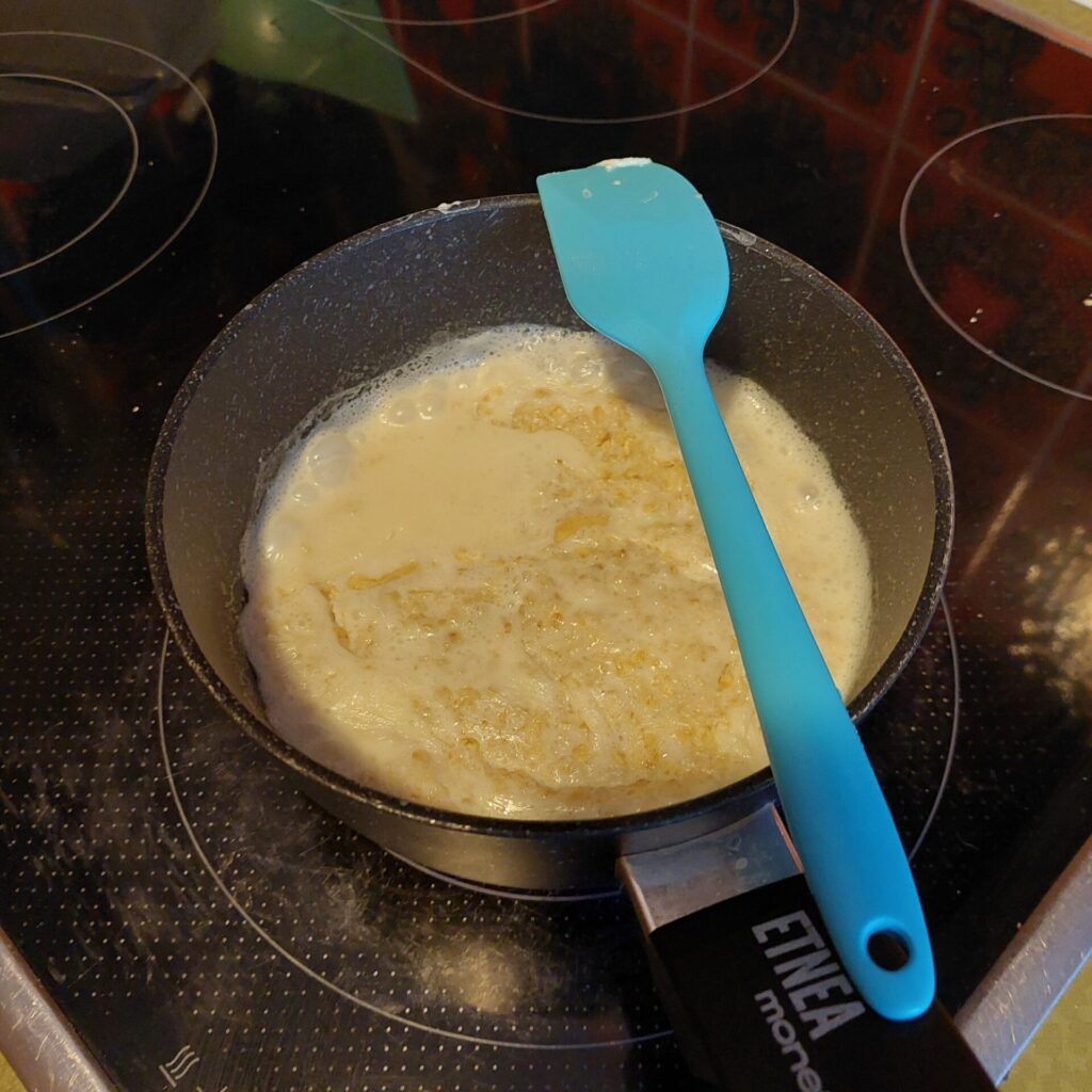 Topf mit Haferflocken und Milch auf einem eingeschalteten Herd. Die Haferflocken werden gekocht, es handelt sich um die Zubereitung eines einfachen Frühstücks.