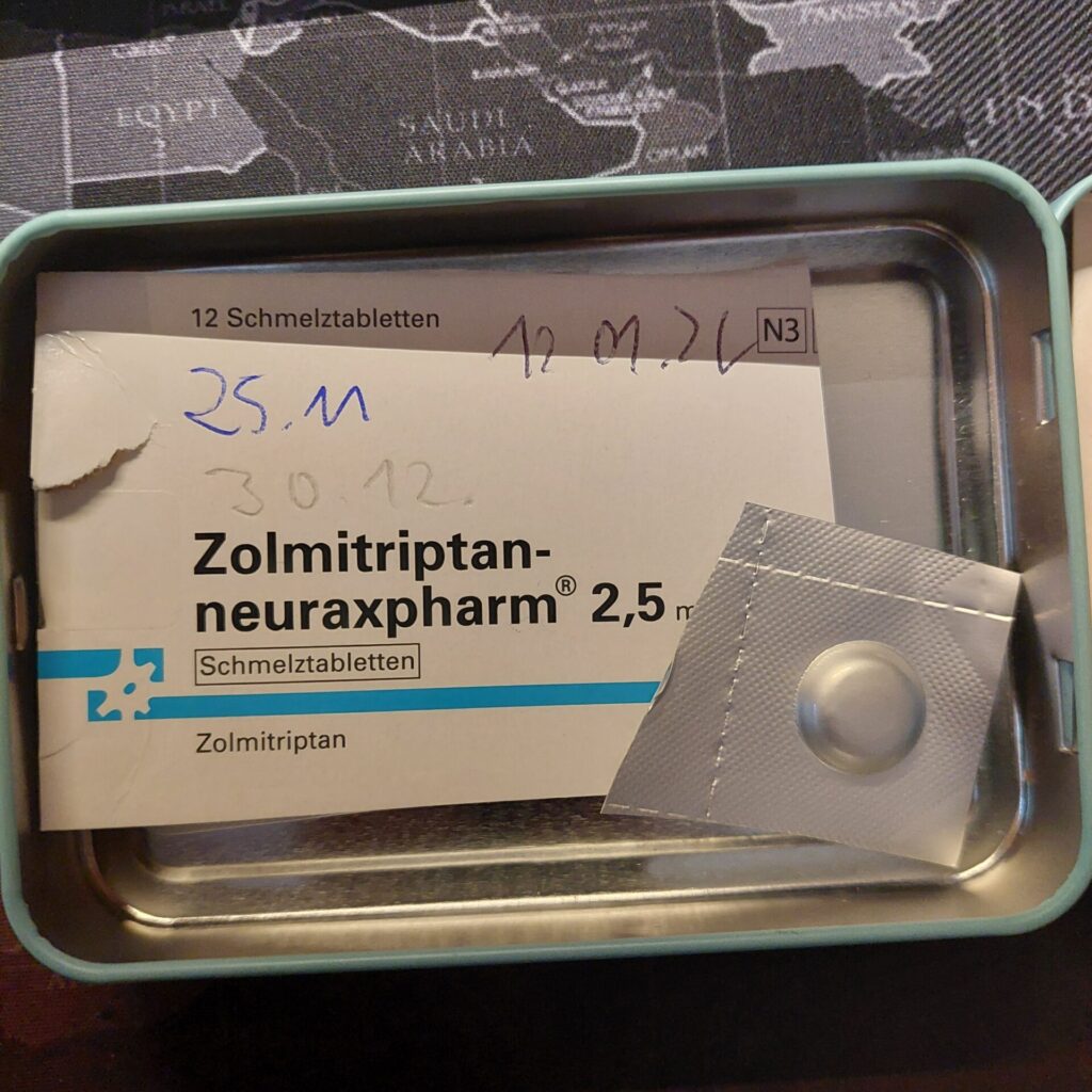 Offene Metallbox mit einer Packung Zolmitriptan-Schmelztabletten (2,5 mg). Daneben liegt eine einzelne Tablette in der Blisterverpackung. Auf der Schachtel sind handschriftliche Datumsnotizen zu sehen.