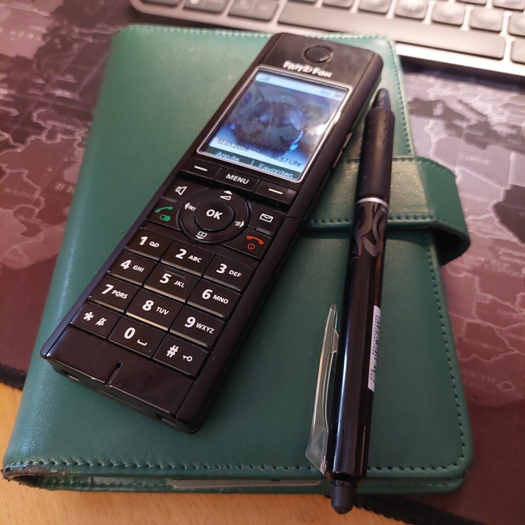 Schwarzes Mobiltelefon mit Tasten liegt auf einem grünen Notizbuch. Daneben befindet sich ein schwarzer Stift. Im Hintergrund ist eine Tastatur zu sehen, die Szene wirkt wie ein Arbeitsplatz.