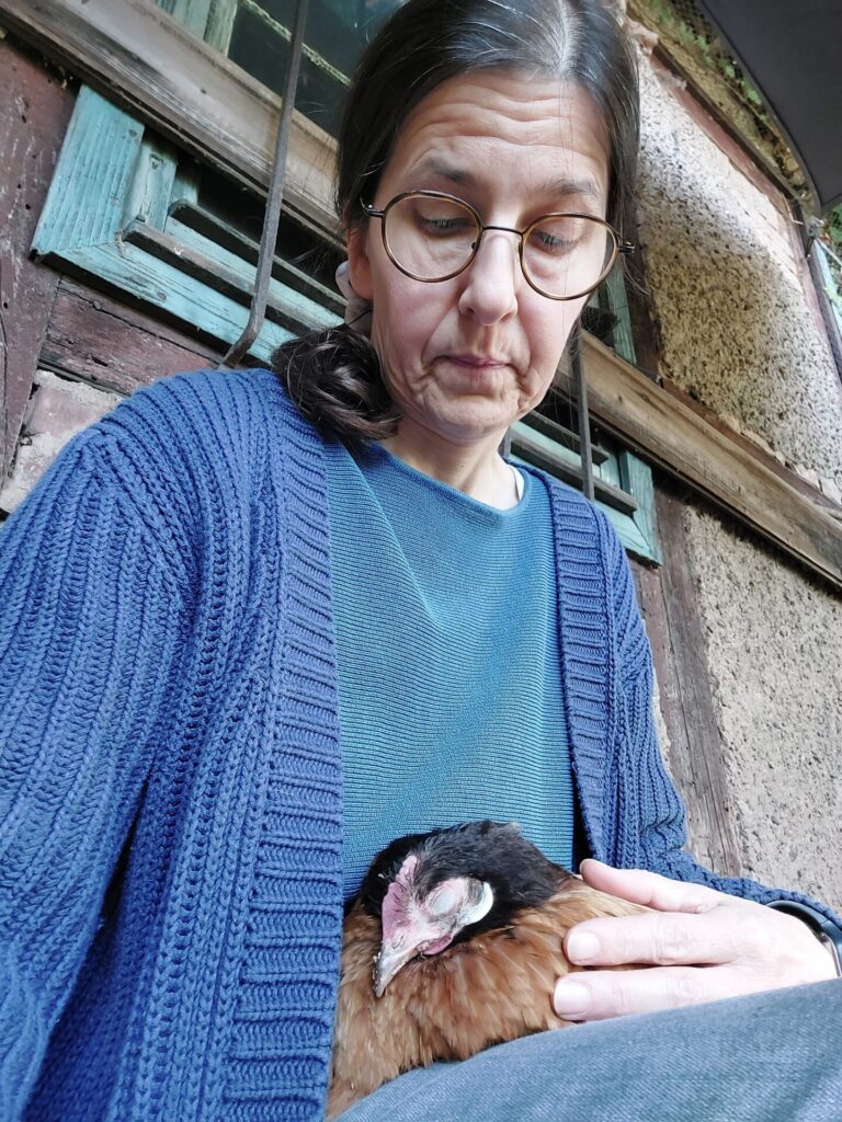 Frau mit Brille und blauem Pullover hält ein verletztes Huhn sanft im Arm. Beide wirken ruhig und verbunden.