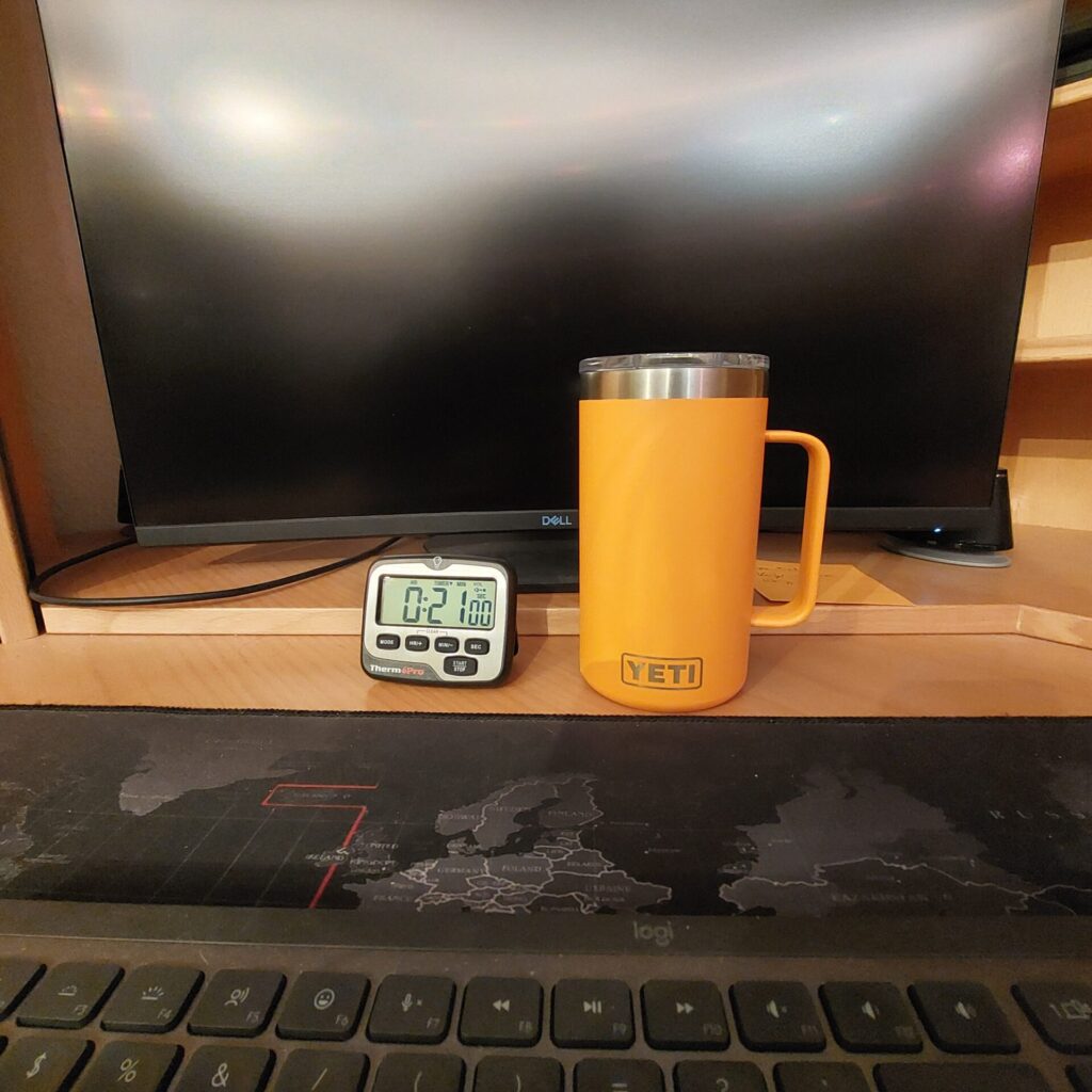 „Schreibtisch am Morgen mit Monitor, Tastatur, orangefarbenem YETI-Becher und einem kleinen digitalen Timer.“