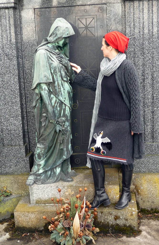 Frau mit Geier-Krafttier-Rock berührt eine Skulptur – Begegnung zwischen Leben, Tod und Transformation