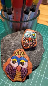 Bemalte Steineulen auf einem Stein arrangiert, im Hintergrund Malstifte