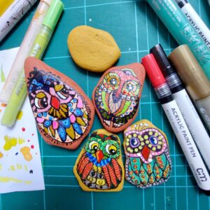 Vier detailreich bemalte Steineulen, umgeben von Farben und Arbeitsmaterial