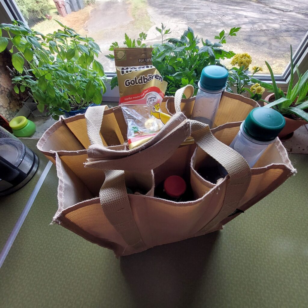 Stofftasche mit verschiedenen Getränkeflaschen und einer kleinen Tüte Gummibärchen steht auf einem Tisch vor einem Fenster mit Kräutertöpfen.