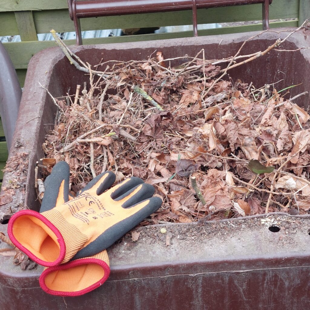 Braune Mülltonne wird mit Gartenabfällen wie Laub und Zweigen gefüllt, Handschuhe liegen daneben.