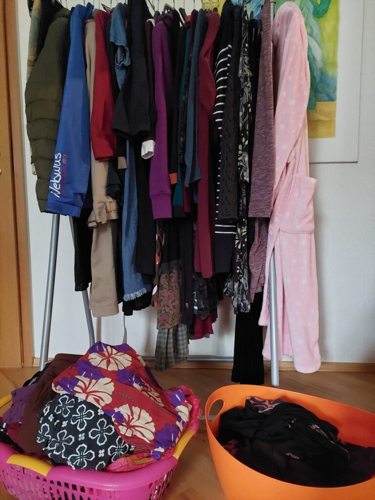 Kleiderständer mit Kleidung in Übergangsphase zwischen Winter und Frühling, Wäschekörbe mit aussortierten und zu sortierenden Kleidungsstücken.