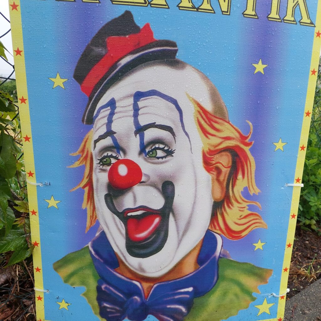 Buntes Schild mit Clownsgesicht, roter Nase und gelben Sternen, auf dem Regentropfen zu sehen sind.