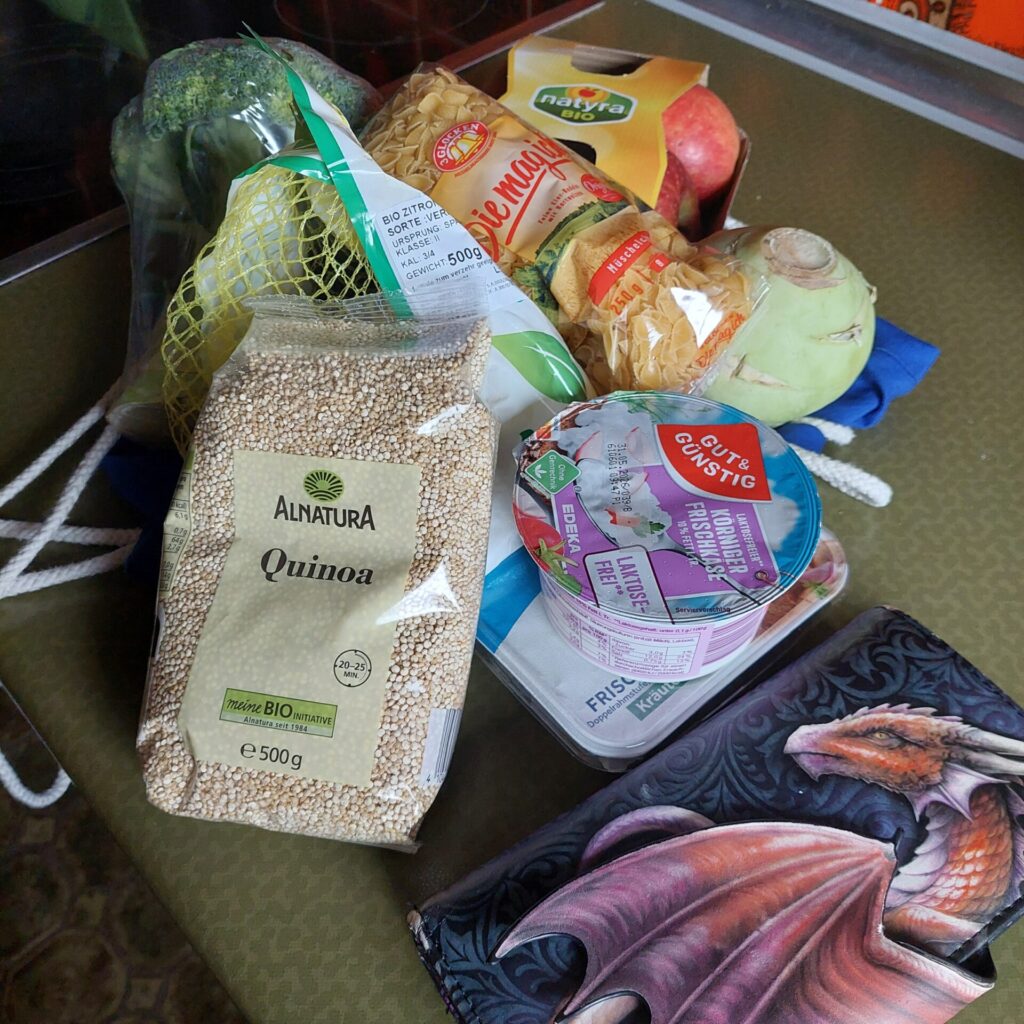 Einkäufe mit Quinoa, Nudeln, Äpfeln, Gemüse und Frischkäse liegen neben einem Portemonnaie mit Drachenmotiv.