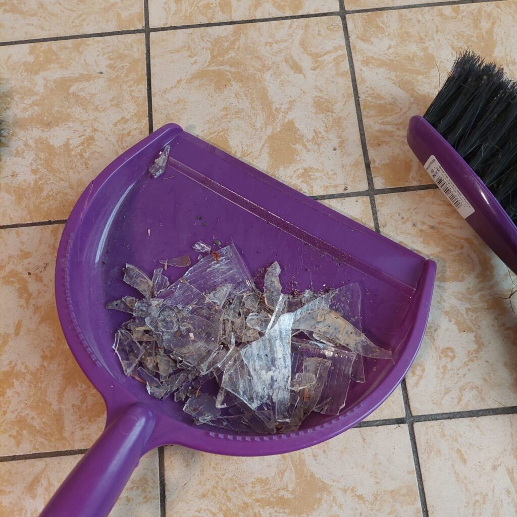 Scherben eines großen Glases für Küchenabfälle liegen auf einer violetten Kehrschaufel neben einem Besen.