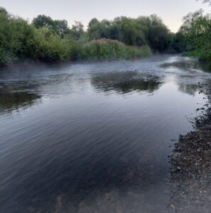 Ruhiger Fluss am frühen Morgen, über dem Wasser liegt leichter Nebel.
