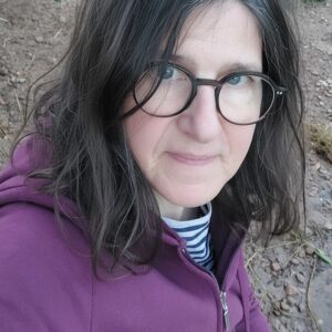Silke mit Brille und weinroter Jacke am Flussufer.