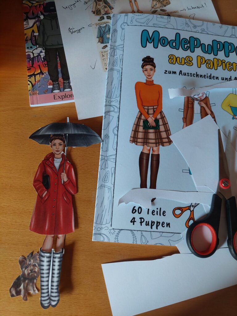 Ausgeschnittene Papierpuppe mit Regenmantel und Hund neben einem Papierpuppenheft, Schere und Papierresten; im Hintergrund liegt der kommentierte Entwurf einer eigenen Gliederpuppe.