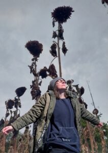Frau in dunkler Kleidung mit Mütze und Brille steht mit ausgebreiteten Armen in einem Feld aus vertrockneten Sonnenblumen unter grauem Himmel.