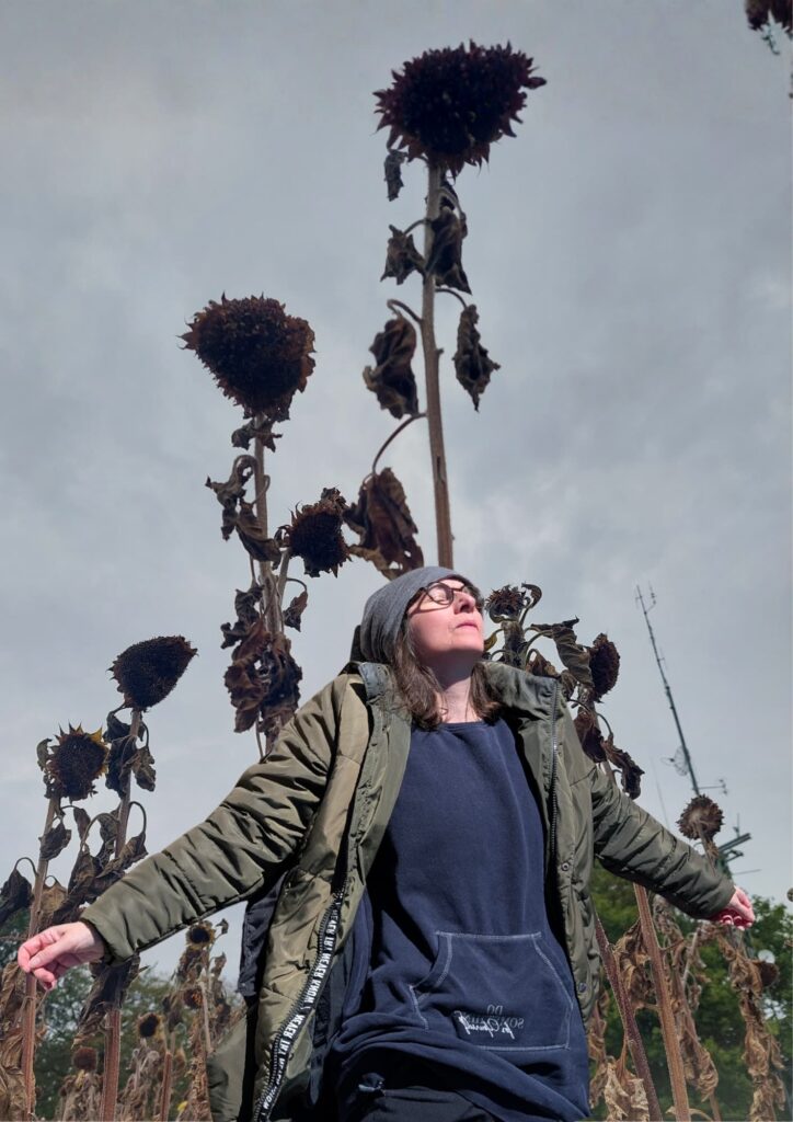 Frau in dunkler Kleidung mit Mütze und Brille steht mit ausgebreiteten Armen in einem Feld aus vertrockneten Sonnenblumen unter grauem Himmel.