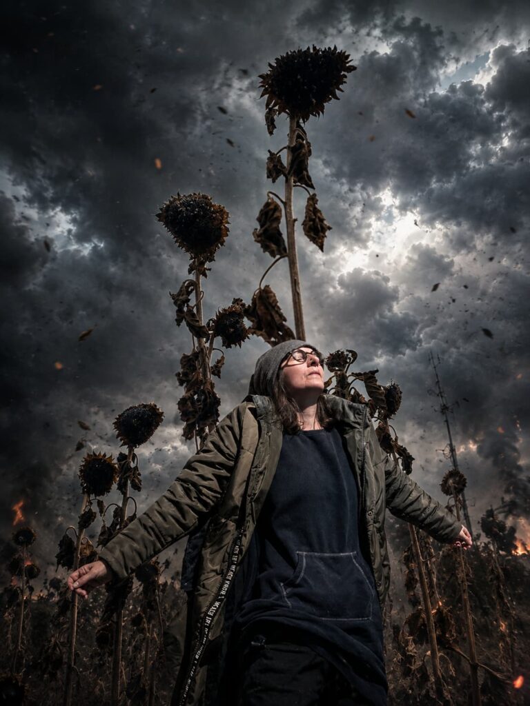 Frau in dunkler Kleidung und Mütze steht mit ausgebreiteten Armen in einem Feld aus vertrockneten Sonnenblumen unter einem düsteren, sturmvollen Himmel.