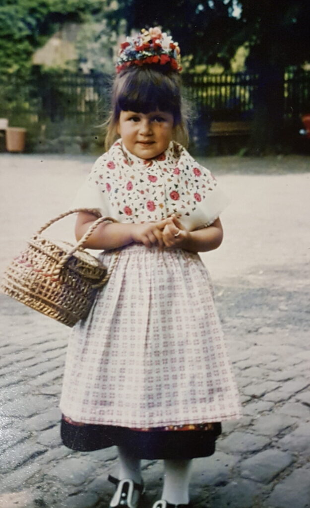 Silke als Kind in festlicher Tracht mit Blumenkopfschmuck neben einem Jungen in Trachtenkleidung; sein Gesicht ist unkenntlich gemacht. Beide stehen vor einer Sandsteinmauer und einem blühenden Garten.