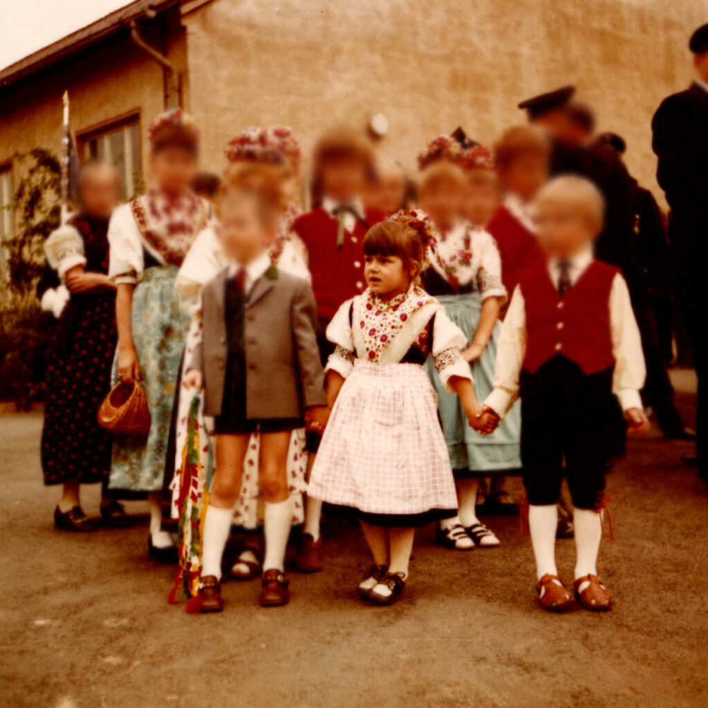 Silke steht 1972 als kleines Mädchen in Tracht im Mittelpunkt einer Kindertrachtengruppe. Sie trägt eine helle Schürze und hält zwei Jungen an den Händen; die Gesichter der anderen Personen sind unkenntlich gemacht.