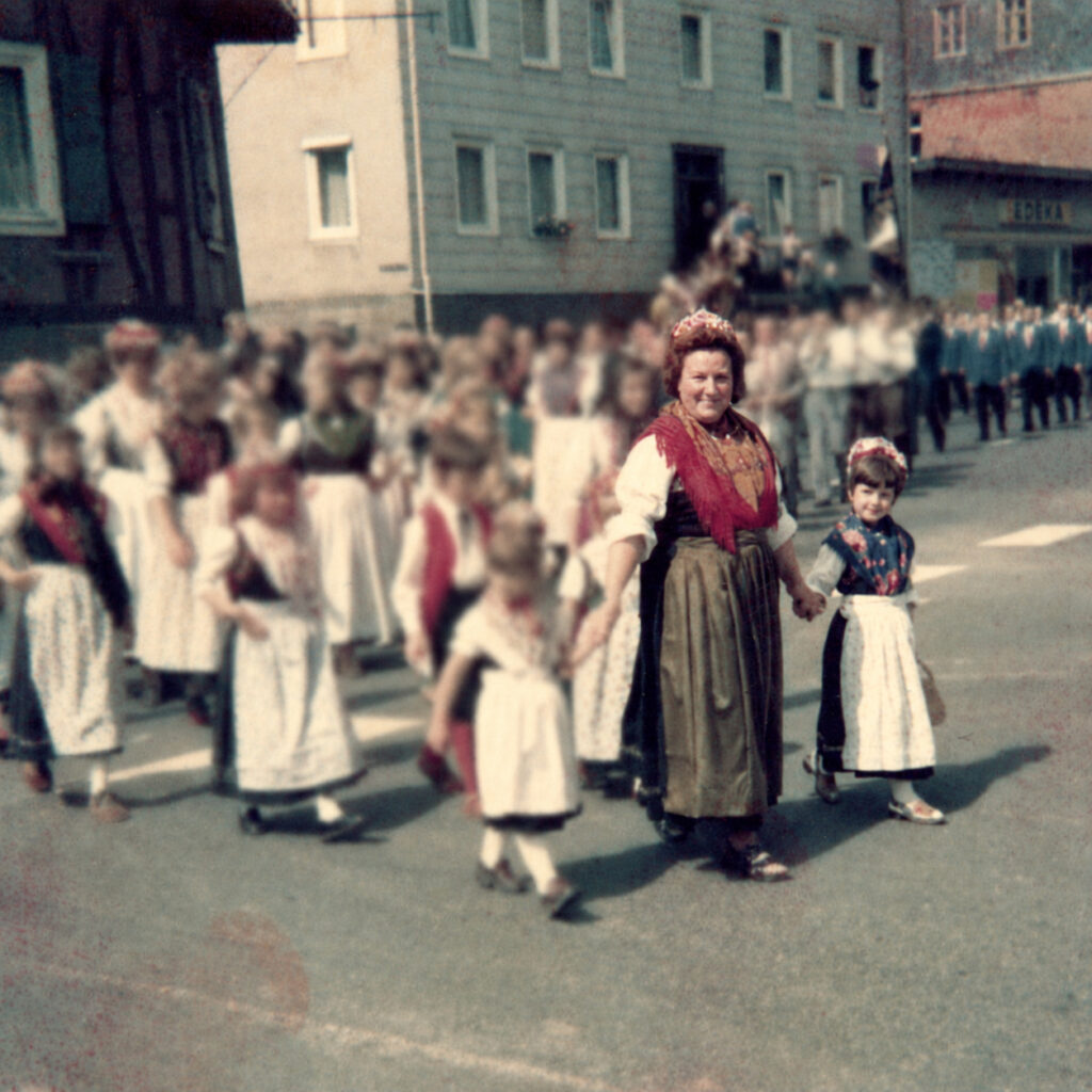 Silke geht als kleines Mädchen in Tracht neben ihrer Mutter bei einem Festumzug durch den Ort. Beide tragen traditionelle Kleidung und halten sich an der Hand; die Gesichter der übrigen Teilnehmenden sind unkenntlich gemacht.