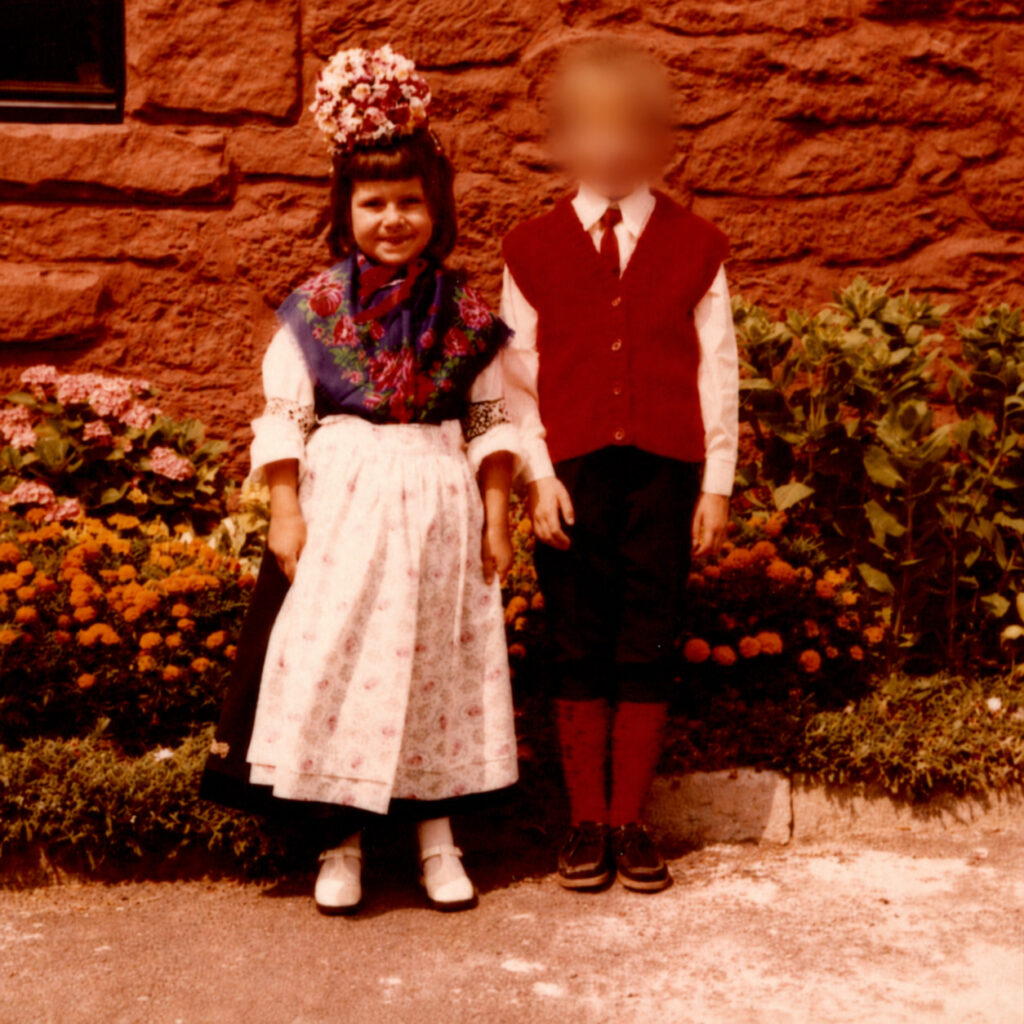 Silke steht als kleines Mädchen in festlicher Tracht mit geblümtem Schultertuch, heller Schürze, Blumenkopfschmuck und geflochtenem Korb auf einem gepflasterten Platz.