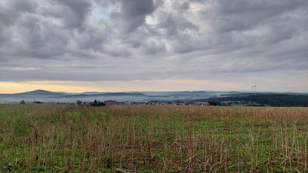 Blick über abgeerntete Felder und eine weite Hügellandschaft unter dramatischem Wolkenhimmel am frühen Morgen.