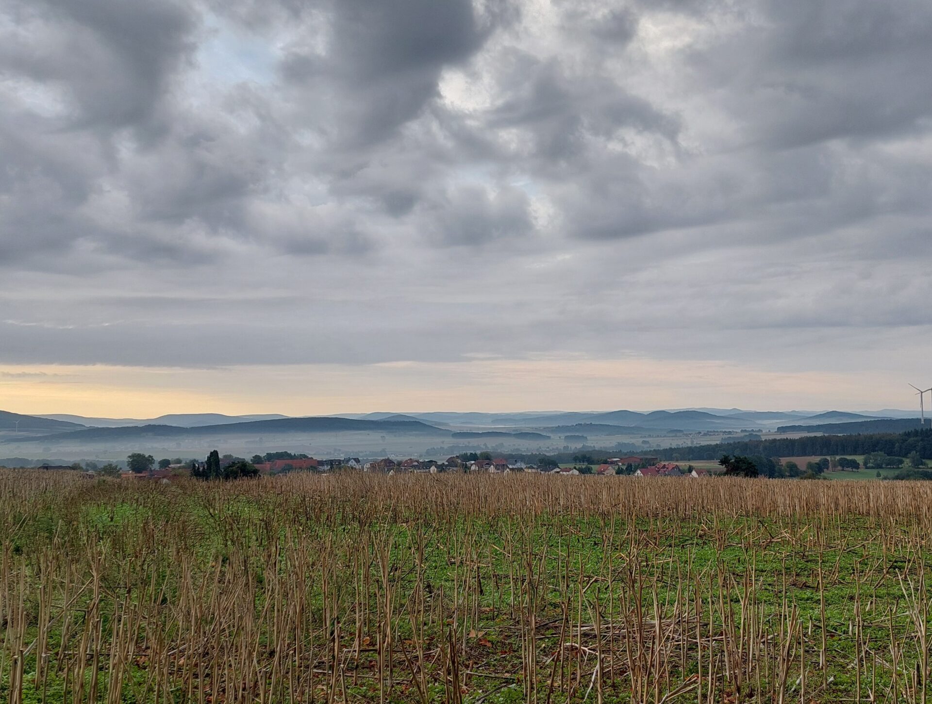 Weite Hügellandschaft mit abgeernteten Feldern unter einem dramatischen Wolkenhimmel am frühen Morgen.